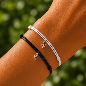 Puede incluir: Dos pulseras de cordón ajustables, una blanca y otra negra, cada una con un pequeño dije plateado en forma de trébol de cuatro hojas. Las pulseras están diseñadas con nudos corredizos simples para el cierre, mostrando un estilo sencillo.