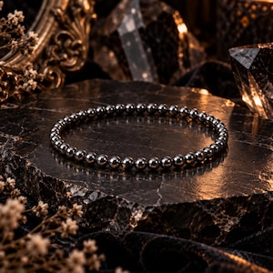 Puede incluir: Una pulsera circular compuesta por pequeñas cuentas esféricas metálicas de color gris oscuro pulido. Las cuentas tienen una superficie lisa y reflectante, ensartadas en un bucle continuo para crear una pieza de joyería uniforme.