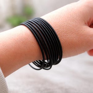 Puede incluir: Un conjunto de pulseras elásticas negras en una muñeca. Las pulseras están hechas de un material fino y flexible y están enrolladas juntas. Las pulseras son de color negro sólido y parecen nuevas.