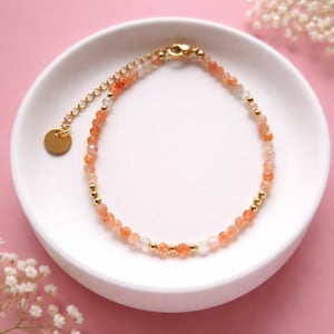 Könnte beinhalten: Ein zartes Armband mit kleinen, facettierten orangefarbenen und klaren Perlen, die mit goldfarbenen Metallperlen durchsetzt sind. Das Armband hat einen goldfarbenen Verschluss und einen kleinen, runden Anhänger. Es wird auf einer weißen, runden Schale vor rosa Hintergrund präsentiert.