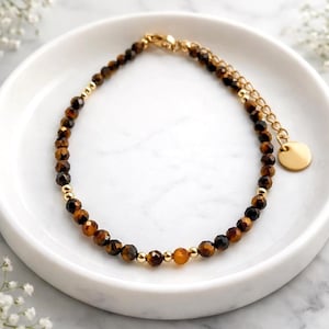 Könnte beinhalten: Ein Armband mit Tigeraugen-Edelsteinen und goldfarbenen Akzenten. Das Armband besteht aus kleinen, facettierten braunen und schwarzen Perlen, die mit goldfarbenen Perlen durchsetzt sind. Eine goldfarbene Kette und ein kleiner, runder Anhänger vervollständigen das Design.