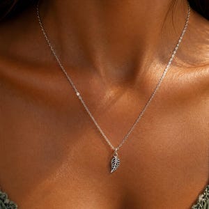 Op de afbeelding: Een verfijnde zilverkleurige ketting met een kleine, gedetailleerde bladvormige hanger met een ingewikkeld uitgesneden patroon. De hanger hangt aan een dunne, eenvoudige schakelketting die op het sleutelbeen rust en een glanzende afwerking heeft.