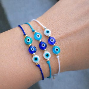 Puede incluir: Primer plano de tres pulseras con cuentas de ojo turco azules y blancas. Cada pulsera tiene un cordón de diferente color: azul oscuro, azul claro y blanco. Las cuentas están acentuadas con cuentas de metal color oro.