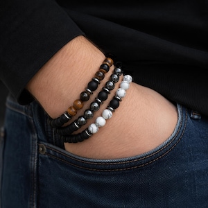 Könnte beinhalten: Drei Armbänder mit Perlen aus Naturstein und Metallakzenten. Die Armbänder weisen eine Mischung aus Farben auf, darunter Braun, Schwarz, Weiß und Silber. Die Armbänder werden am Handgelenk getragen, mit einem schwarzen Hemd und einer blauen Jeans.