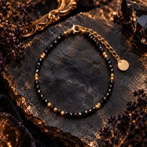 Op de afbeelding: Een armband met kleine, gefacetteerde zwarte kralen afgewisseld met gepolijste goudkleurige metalen bolletjes. De armband is voorzien van een goudkleurige karabijnsluiting, een verstelbare verlengketting en een kleine, ronde, blanco metalen bedel.