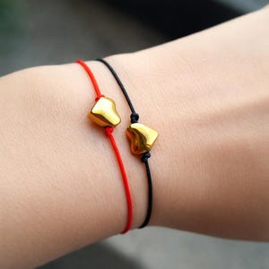May include: Two matching bracelets. One has a red cord and the other has a black cord. Each bracelet features a gold-colored, heart-shaped charm. The bracelets are worn on a wrist.