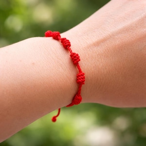 Pode incluir: Uma pulseira vermelha brilhante com nós. A pulseira é feita de um cordão vermelho fino com vários nós espaçados uniformemente. A pulseira é usada em um pulso, com um fundo verde desfocado.