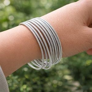 Op de afbeelding: Een set zilverkleurige, opgerolde draadarmbanden die om een pols worden gedragen. De armbanden zijn gemaakt van dun, flexibel metaal en hebben een glanzend, reflecterend oppervlak. De achtergrond is zachtgroen, wat duidt op een buitenomgeving. Een elegant accessoire.