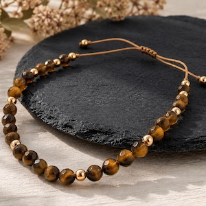 Könnte beinhalten: Ein Armband mit facettierten braunen Tigeraugen-Perlen, die sich mit glatten goldfarbenen Metallperlen abwechseln. Das Schmuckstück verfügt über eine beigefarbene Kordel mit verstellbarem Schiebeknoten und kleinen goldfarbenen Perlen an den Enden.