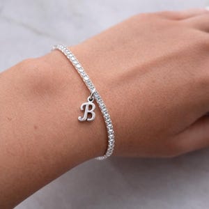 Può includere: Un braccialetto d'argento con una fila di piccole pietre trasparenti. Un ciondolo d'argento a forma di lettera "B" pende dal braccialetto. Il braccialetto è indossato su un polso.