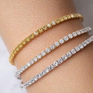 Op de afbeelding: Drie tennisarmbanden. Eén is goudkleurig met gele stenen, de andere twee zijn zilverkleurig met heldere stenen. De armbanden worden om een pols gedragen, wat hun elegante ontwerp en glans benadrukt.