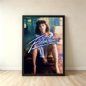 May include: A framed poster featuring the movie title "Flashdance" in a stylized blue font. The image shows a person in a blue top and red heels. The background has a blurred, colorful effect, with a vintage aesthetic.