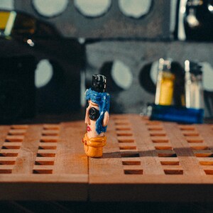 May include: A quirky, hand-painted figurine with a blue wig, black hat, and button eyes. The figurine has a yellow base and red lips, standing on a wooden surface. The background includes industrial-style concrete blocks and other objects.