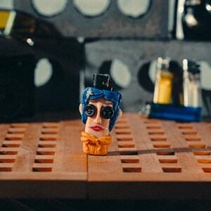 May include: A quirky, hand-painted figurine of a character with blue hair, large button eyes, and a small black hat. The figurine has a yellow scarf and red lips. It is displayed on a wooden surface with a grid pattern.