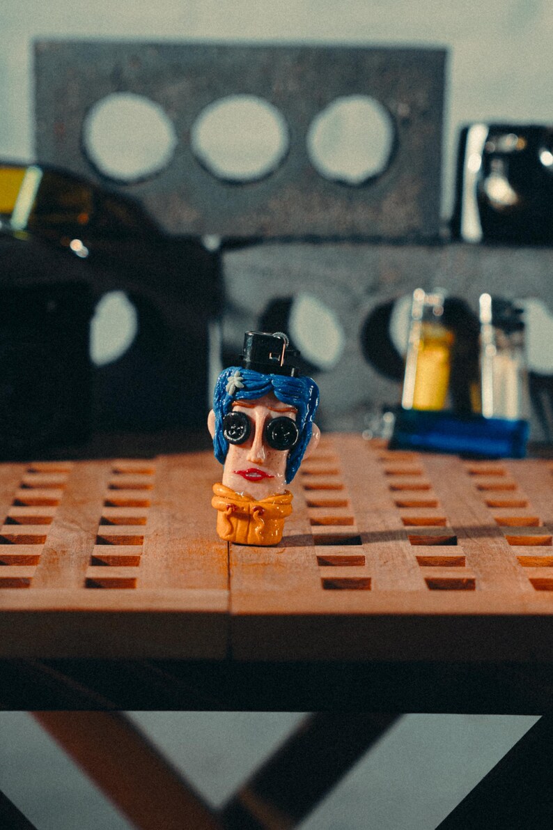 May include: A quirky, hand-painted figurine of a character with blue hair, large button eyes, and a small black hat. The figurine has a yellow scarf and red lips. It is displayed on a wooden surface with a grid pattern.