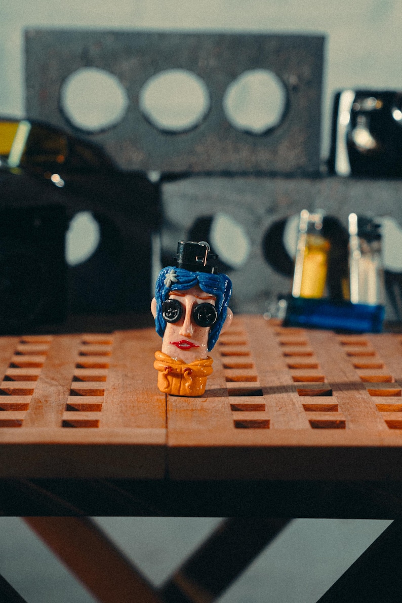 May include: A quirky, hand-painted figurine with a blue-haired head, button eyes, and a small black hat. The figure has a yellow scarf and red lips. It sits on a wooden surface with a grid pattern, with a blurred background.