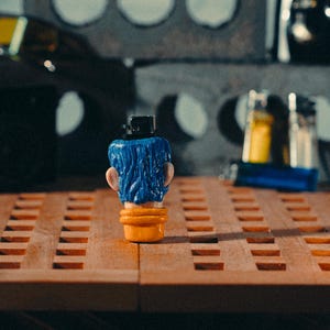 May include: A novelty lighter in the shape of a head with blue hair, a black hat, and an orange base. The lighter is on a wooden surface with a grid pattern. Other objects are visible in the background, including a concrete block.