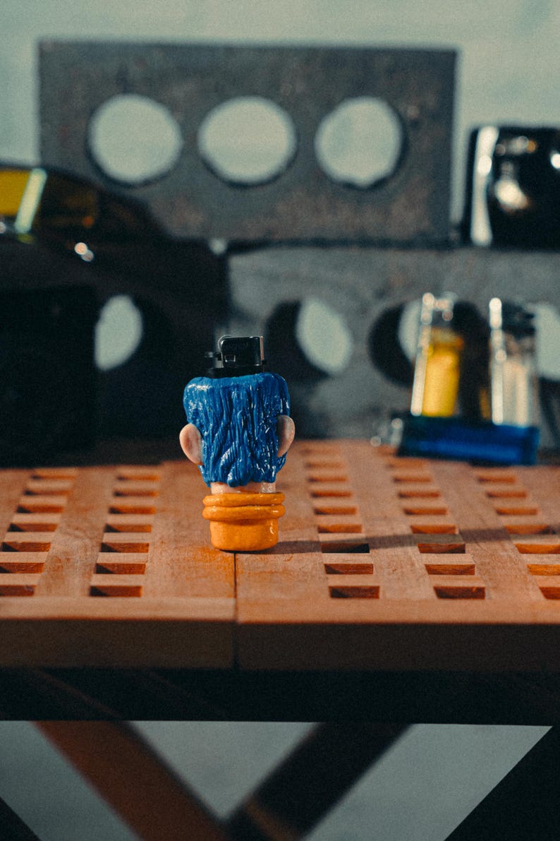 May include: A novelty lighter in the shape of a head with blue hair, a black hat, and an orange base. The lighter is on a wooden surface with a grid pattern. Other objects are visible in the background, including a concrete block.
