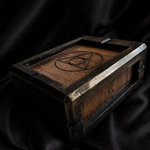Peut inclure: Une boîte rectangulaire en bois avec une finition marron foncé et des bordures métalliques. Le couvercle présente un motif géométrique gravé. La boîte a un aspect vintage et artisanal, idéale pour ranger de petits objets.