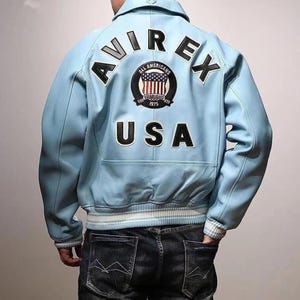 May include: Light blue leather jacket with the word "AVIREX" across the shoulders and "USA" on the lower back. A circular patch with the American flag and the words "ALL AMERICANS ESTABLISHED 1975" is centered on the back.