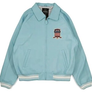 May include: A light blue jacket with a collared neckline and a zipper closure. The jacket features a patch with an American flag design and the brand name. The cuffs and hem have white and light blue striped detailing.