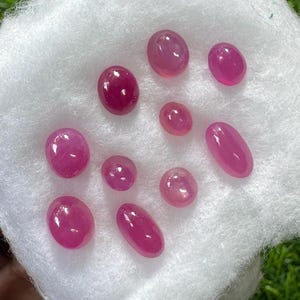 Puede incluir: Una colección de nueve piedras preciosas rosas, lisas y de forma ovalada, dispuestas sobre un algodón blanco. Las piedras varían en tamaño y presentan un aspecto pulido y translúcido con tonos magenta y rosa claro que reflejan la luz.