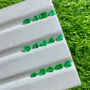 May include: A collection of small, faceted green gemstones arranged in three horizontal rows on a white, textured display tray. The stones feature various cuts, including oval, marquise, square, and rectangular shapes, all displaying a vibrant green hue.