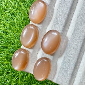May include: Five smooth, oval-shaped peach-colored moonstone cabochons with a polished finish. Each gemstone displays a soft, translucent glow and a rounded surface, arranged in a cluster on a textured, off-white display stand.