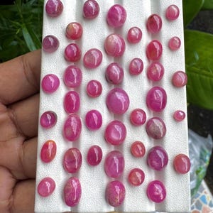 May include: A collection of numerous small, polished, oval and round pink gemstones arranged in rows on a white, ridged display tray. The stones vary in size and exhibit a smooth, glossy surface with translucent and opaque pink color variations.