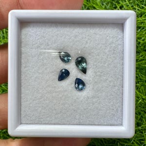 Könnte beinhalten: Vier birnenförmige, lose Edelsteine in Blau- und Petroltönen befinden sich in einem quadratischen weißen Kunststoffetui mit transparentem Deckel. Die facettierten Steine sind auf einer weißen Filzeinlage angeordnet und reflektieren das Licht.