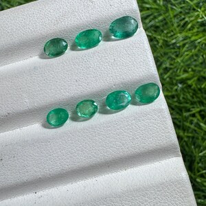 Peut inclure: Sept pierres précieuses émeraude de taille ovale avec une teinte verte vibrante et des surfaces facettées. Les pierres sont disposées en deux rangées sur un présentoir blanc texturé, mettant en valeur leur qualité translucide et leur fini poli.