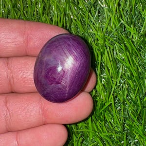 Op de afbeelding: Een gladde, ovaalvormige paarse edelsteen met een zichtbaar zeszalig sterpatroon op het oppervlak. De steen vertoont diepe magenta en violette tinten met een gepolijste, reflecterende afwerking die een natuurlijk asterisme-effect laat zien.