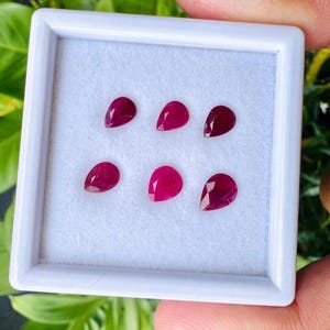 May include: Six loose, pear-shaped rubies with a deep red color and faceted surfaces, arranged in two rows of three inside a square white display box with a soft white velvet-like insert. The gemstones exhibit clear, reflective surfaces.