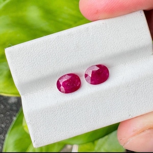 May include: Two oval-cut, deep red rubies displayed on a white textured display card. The gemstones feature a faceted surface that reflects light, showcasing a vibrant crimson hue. The stones are positioned side-by-side on the white material.
