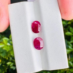 Pode incluir: Dois rubis de corte oval e vermelho profundo são exibidos em um cartão de apresentação branco e texturizado. As pedras preciosas apresentam superfícies facetadas que refletem a luz, destacando sua cor carmesim vibrante e brilho polido.
