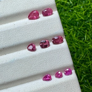 Pode incluir: Uma coleção de sete pedras preciosas cor-de-rosa facetadas dispostas num expositor branco com três ranhuras horizontais. As pedras variam na forma, incluindo cortes em pera e ovais, e exibem uma cor magenta-rosa vibrante e translúcida.