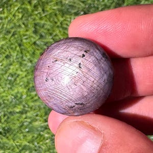 May include: A round, cabochon-cut star ruby gemstone with a dusty mauve and purple hue. The surface displays a visible six-rayed star effect, natural inclusions, and fine surface striations across the polished, domed top of the stone.