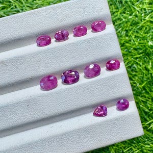 Peut inclure: Une collection de dix rubis roses à facettes disposés sur un présentoir blanc. Les pierres précieuses sont principalement de forme ovale, avec une pierre en forme de poire incluse. Chaque rubis présente une teinte rose vive et translucide.