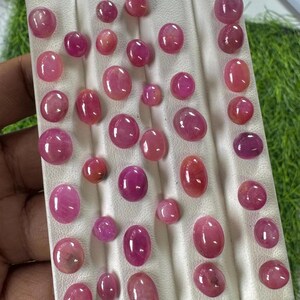 May include: A collection of numerous oval and round cabochon-cut pink and reddish-pink gemstones arranged in rows on a white display board. The stones exhibit varying shades of pink, ranging from light rose to deep magenta, with smooth surfaces.
