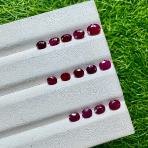 Pode incluir: Uma coleção de catorze rubis facetados de corte oval dispostos em três filas num expositor branco. Cada pedra preciosa apresenta uma cor vermelha profunda e uma superfície polida, realçando a clareza e o brilho característicos dos rubis.