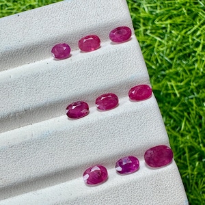 May include: Eight oval-cut, deep pinkish-red rubies arranged in three rows on a white, textured display stand. Each gemstone features a faceted surface that reflects light, showcasing a consistent color and shape across the collection of loose stones.