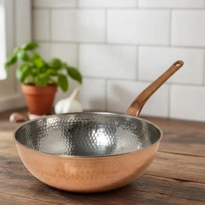 Può includere: Una padella in rame tipo wok con interno martellato e un lungo manico color rame. La padella è su una superficie di legno, con uno scorcio di una pianta in vaso e aglio sullo sfondo. Il design della padella suggerisce che è per cucinare.