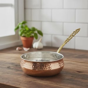 Puede incluir: Una cacerola de color cobre con acabado martillado y asa de latón. El interior es plateado. La cacerola está sobre una superficie de madera, con una pequeña planta en maceta, ajo y un salpicadero de azulejos blancos al fondo.