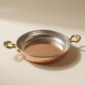 May include: A copper-toned, round pan with a hammered finish and two decorative gold-coloured handles. The pan has a shiny interior and a slightly raised rim. The handles are curved and have a detailed design. Likely used for cooking or serving.