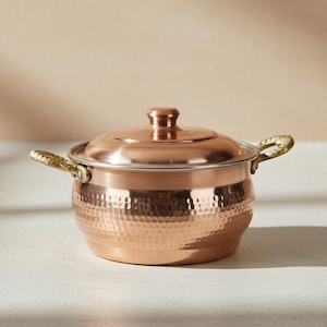May include: A copper-colored cooking pot with a lid and two handles. The pot has a hammered texture on the body and a shiny finish. The handles are gold-colored. The pot is sitting on a white surface.
