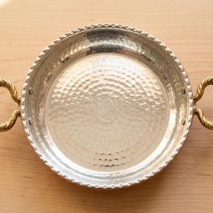 May include: A silver-colored metal serving tray with a hammered texture and two gold-colored rope-style handles. The rim has a decorative beaded edge. The tray is round and sits on a light brown wooden surface.