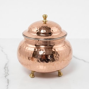 May include: A copper-coloured, hammered metal jar with a lid and three small feet. The jar has a rounded body and a lid with a small, decorative knob. The metal has a shiny, reflective surface.