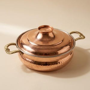 May include: A copper cooking pot with a lid. The pot has a hammered texture on the lower portion and a smooth, reflective surface on the lid. It features brass handles and a matching brass knob on the lid. The pot is designed for cooking.