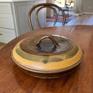 May include: A round, lidded ceramic casserole dish featuring a glossy, dark brown center and a wide, textured band of mustard yellow and olive green glaze. The lid includes a curved, arched handle, and the base has small side handles.