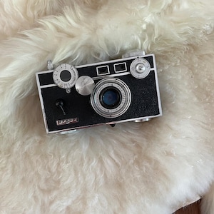 May include: A vintage Argus rangefinder camera with a black textured body and silver metal accents. The front features a circular lens, a rangefinder dial, a shutter speed dial, and a small black lever. The word Argus is printed in white.